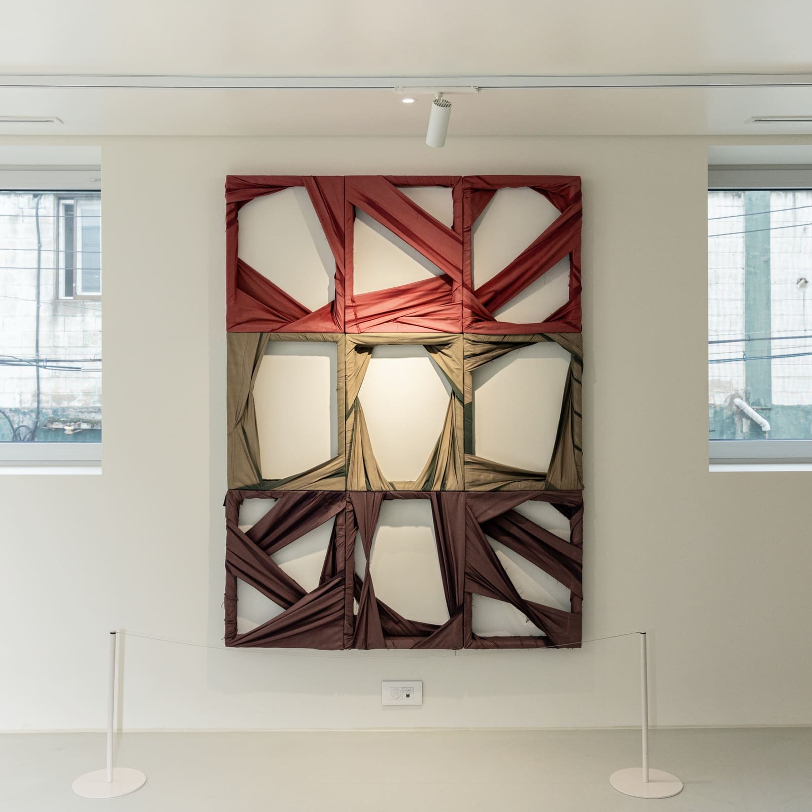 Nine frames wrapped in red, khaki and brown fabric, hung on a white gallery wall